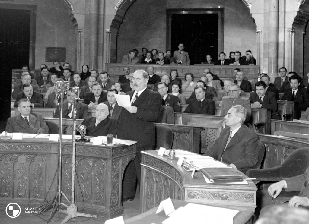 Nagy Imre (k), mint a Minisztertanács elnöke 1954. január 23-án felszólal az Országgyűlésen. Mellette Rákosi Mátyás (b2) a Magyar Dolgozók Pártjának (MDP) első titkára és Gerő Ernő (j) a Minisztertanács első elnökhelyettese, belügyminiszter ül. Fotó: Nemzeti Archívum